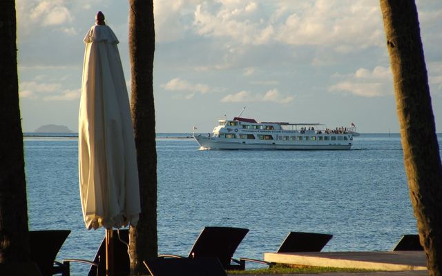 Wit passagiersschip vaart uit vanaf Denarau, gezien vanaf een strand met palmen en ligbedden